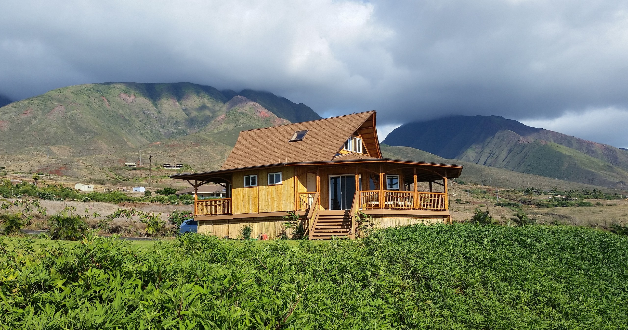 Wooden house with porch in green field, mountains in background under cloudy sky.